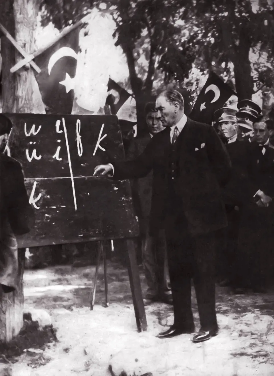 Açık havada, bayrakla örtülü bir kürsünün yanında kara tahtaya yazılmış yeni Türk harflerini gösteren Mustafa Kemal; çevresinde dinleyenler.