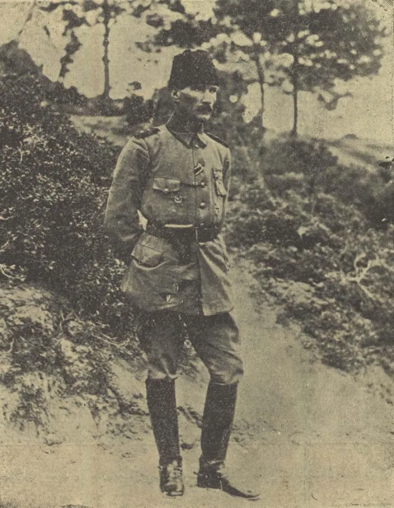 Kabalaklı ve üniformalı Mustafa Kemal Bey, ağaçlık bir yamaçta ayakta duruyor.