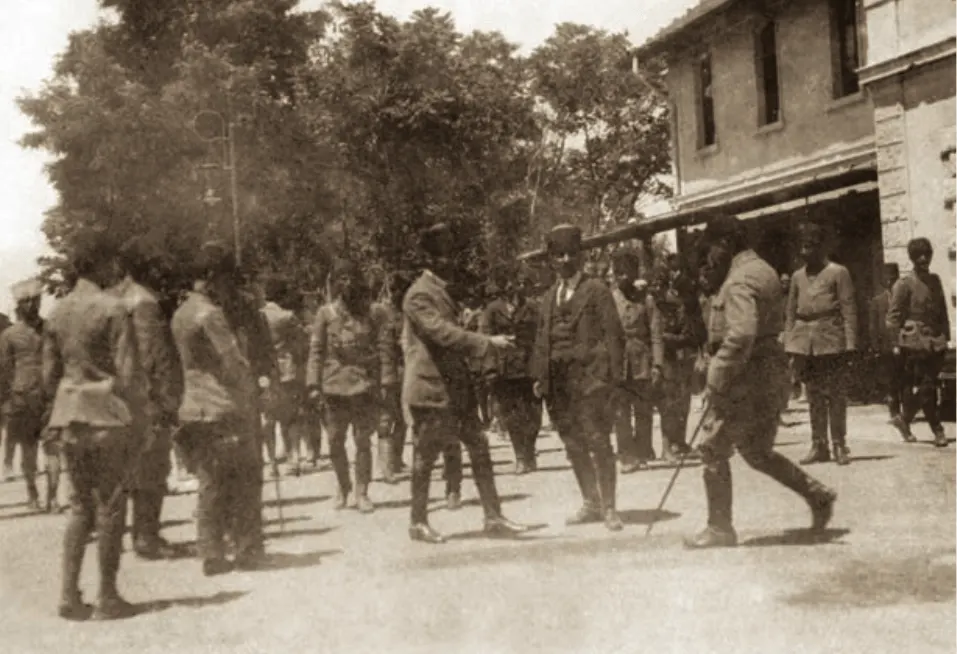 Ağaçlı bir sokakta subaylar arasında yürüyen Mustafa Kemal Paşa; elinde baston var.