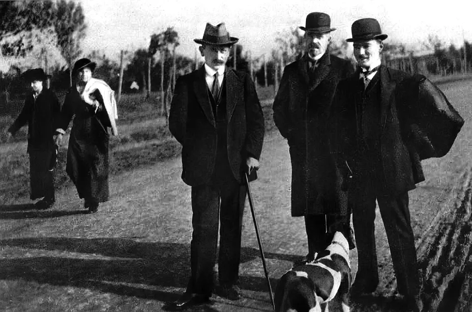 Ağaçlı açık bir arazide melon şapkalı ve paltolu beş erkek. Öndeki üçü kameraya bakıyor; sağ alttan bir av köpeği kadraja giriyor.