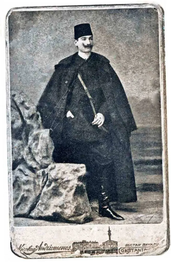 Pelerinli ve fesli Kurmay Yüzbaşı Mustafa Kemal, bir stüdyo dekorunun önünde ayakta; kartonun alt kenarında fotoğrafhanenin damgası var.