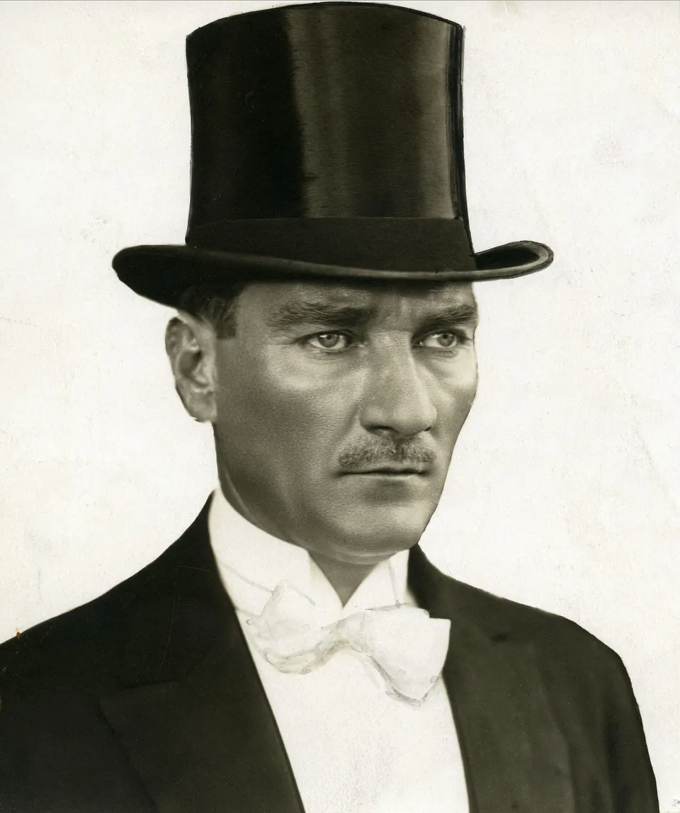 Silindir şapka, beyaz kravat ve frak ceketiyle ayakta duran Mustafa Kemal; bir elinde şapka, diğerinde baston.