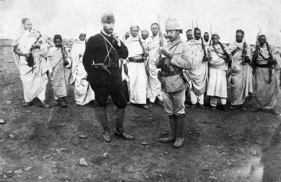 Açık ve taşlık bir arazide, yerel kıyafetli ve silahlı bir grubun önünde duran iki subay. Biri koyu üniformalı ve kalpaklı, diğeri açık renk üniforma ve kolonyal şapkayla bir kâğıda bakıyor.