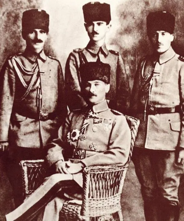 Hasır koltukta oturan, madalyalı ve kalpaklı Mustafa Kemal Paşa; arkasında üniformalı üç subay ayakta.